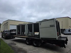 Large HVAC units loaded on a flatbed trailer hitched to a black pickup truck outside industrial buildings under cloudy sky.