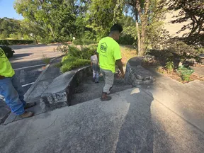 Workers in neon shirts inspecting stone steps and greenery near a parking lot on a sunny day