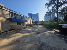 Urban outdoor scene with a beige building, concrete walkway, greenery, parked black truck, and glass office buildings in background.