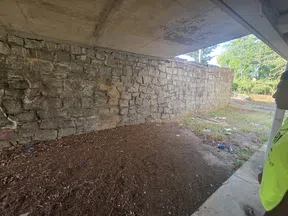 Underbridge area with a stone retaining wall, dirt ground, scattered trash, and a person in a neon shirt.