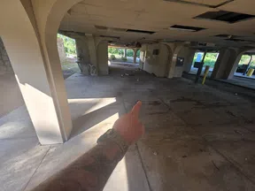 Person pointing inside an empty, abandoned concrete building with arches and scattered debris on the floor.