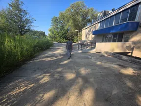 Person walking on a sunny concrete path beside a building with blue awning and green trees in background.