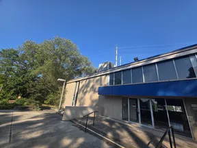 Exterior view of a modern building with large windows, blue awning, and surrounding trees under a clear blue sky.