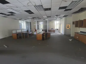 Large empty office space with damaged ceiling tiles, scattered debris, desks, and chairs under fluorescent lighting.