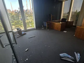 Empty office room with overturned chair, scattered debris, wooden desks, and large windows letting in natural light.