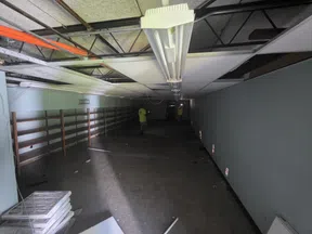 Dimly lit, empty room under renovation with ceiling panels removed and workers in yellow shirts.