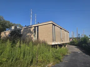 Abandoned brick building with overgrown weeds and cracked pavement under a clear blue sky.