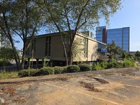 Mid-century modern office building surrounded by trees with a glass high-rise in the background on a sunny day.
