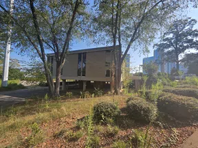 Modern building elevated on stilts surrounded by trees and greenery with city skyscrapers in the background under clear sky