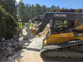 Yellow John Deere compact track loader breaking concrete with hydraulic breaker attachment on construction site.