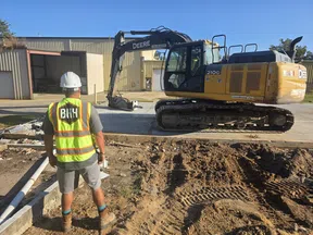 Construction worker in safety vest and helmet overseeing John Deere excavator at a building site with dirt and tire tracks.