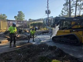 Construction workers in safety gear operating a John Deere skid steer with a hydraulic hammer on concrete.