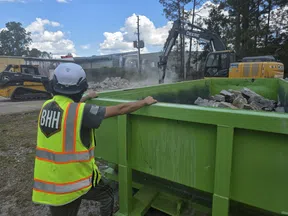 Construction worker in safety gear overseeing debris being loaded into a green dumpster with heavy machinery in background