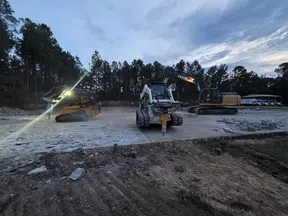 Heavy construction machinery including excavators and a skid steer loader working on a site at dusk with pine trees in the background