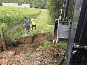 Two men inspect a large washed-out dirt and gravel driveway with an excavator bucket nearby in a rural area.