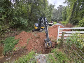 Small excavator digging a trench near a fence and wooded area for construction or drainage work.
