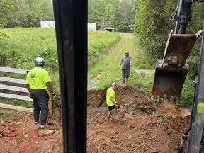 Construction workers in neon shirts excavate soil on a rural dirt road near a white fence and greenery.