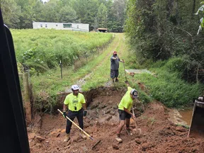 Three workers in safety gear digging and clearing a muddy ditch near a grassy hillside by a house.
