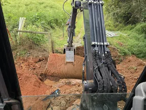 Excavator installing a large corrugated metal pipe in a muddy trench surrounded by green vegetation.