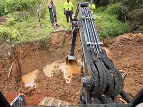 Excavator digging a large muddy hole with workers standing nearby on a grassy hillside.