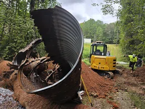 Large rusted metal culvert pipe removed from a trench with a yellow mini excavator and workers in a wooded area.
