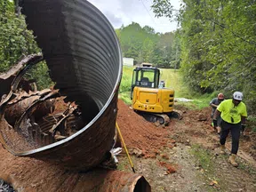 Workers with excavator removing large rusted metal pipe from forested dirt road construction site