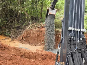 Excavator pouring gravel into a construction site surrounded by trees and red soil.