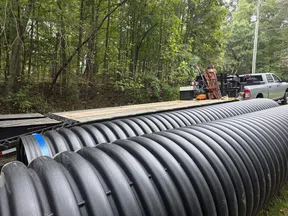 Large black corrugated drainage pipes stacked near a pickup truck and forested area ready for installation.