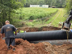 Workers installing large black corrugated drainage pipe in a rural area near a mobile home and dirt road.