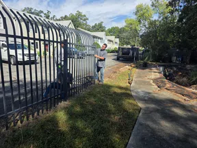 Two men installing a black metal fence along a residential street on a sunny day with parked cars and houses.