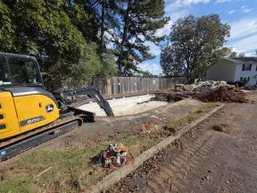 Excavator demolishing an empty swimming pool in a residential backyard under clear sky.