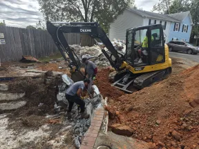 Workers using excavator and tools to demolish concrete structure near residential area with dirt mound.