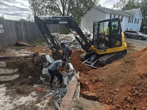 Construction workers operate a John Deere excavator breaking up concrete near a residential house.