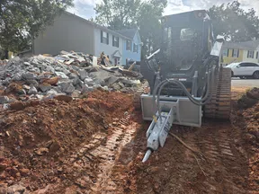 Excavator with hydraulic breaker demolishing ground near rubble pile in residential area with houses.