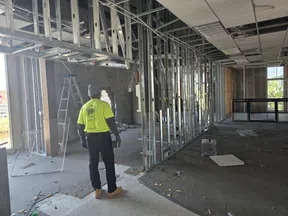 Construction worker in neon shirt stands inside partially built interior with metal framing and debris on floor.