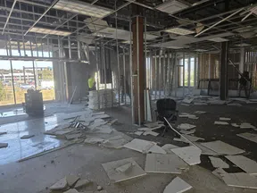Construction site interior with debris, metal framing, and workers installing drywall panels.