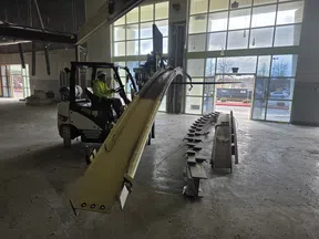 Worker operating forklift lifting a large curved metal beam inside a spacious industrial warehouse.