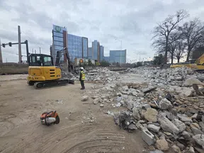 Construction site with worker, excavators, and large piles of rubble near city buildings on a cloudy day