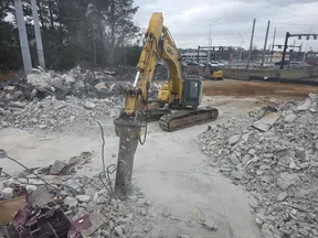 Heavy excavator with hydraulic breaker demolishing concrete rubble on a construction site near a busy road.