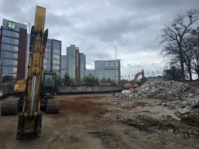 Construction site with excavators, rubble piles, and office buildings under cloudy sky at dusk.