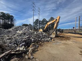 Construction site with excavator clearing large piles of rubble and debris under a blue sky with power lines.