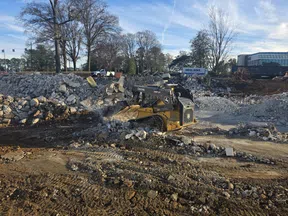 Yellow John Deere excavator moving rubble at a construction site under a blue sky with trees and buildings nearby