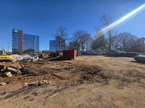 Construction site with dirt ground, machinery, a red dumpster, and modern glass buildings under clear blue sky.