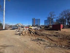 Construction site with excavators, rubble piles, and modern office buildings in the background under clear blue sky