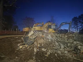 Excavators work on a demolition site with rubble and debris under a twilight sky with trees in the background.