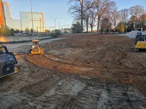 Construction site with earthmoving vehicles leveling soil near office buildings and trees in late afternoon light