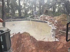 Partially demolished concrete swimming pool with construction machinery and debris around in outdoor setting.