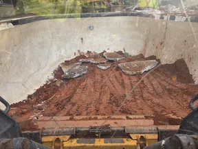 Excavator bucket filled with red soil and broken concrete pieces during a digging operation.