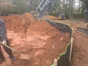 Construction site with excavator digging red soil and worker holding a shovel near erosion control barrier.