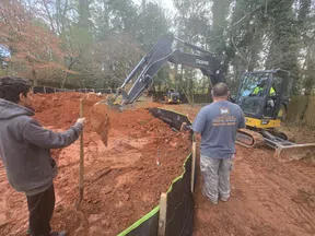 Construction workers operating excavator to dig red soil with silt fence and wooded background on a clear day